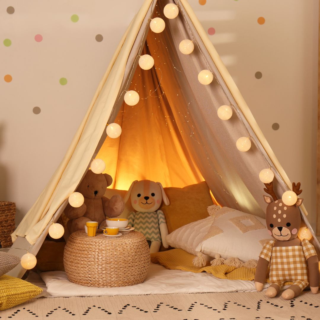 Barnerom, lyst telt pyntet med lyslenke, omgitt av myke puter, kosedyr og lekeservise på gulvet.