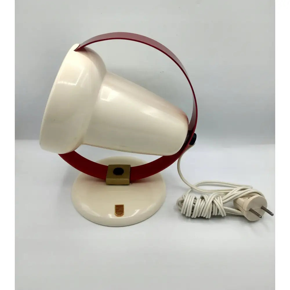 Spotter, sylinderformet hvit lampe med rødt stativ og rund base, hvit ledning og støpsel til høyre.