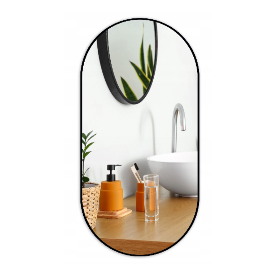 Speil, rundt speil med svart ramme henger over servant og benk med såpedispenser, glass, kurv og grønn plante.