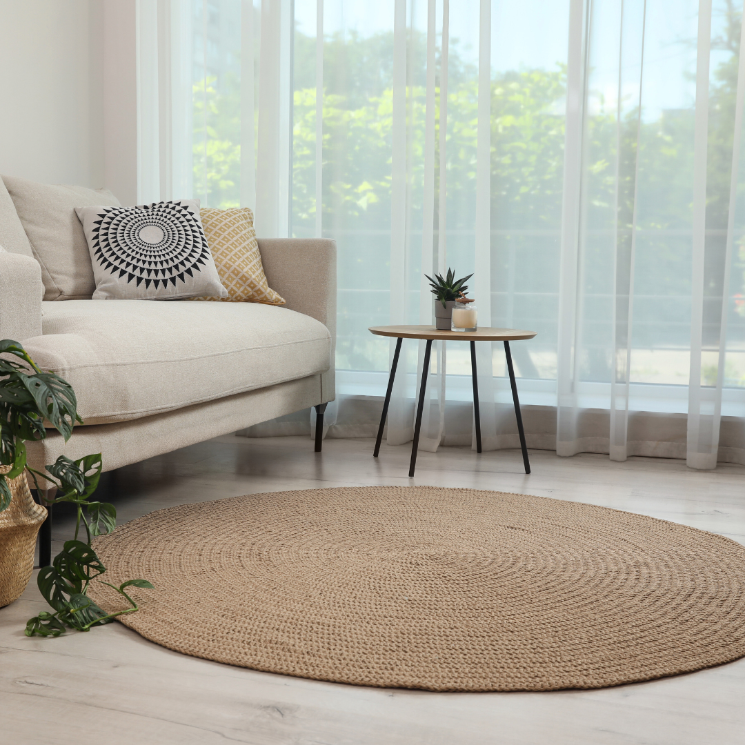 Tepper, beige rundt teppe plassert på lyst tregulv foran lys sofa med puter, lite bord og grønne planter ved siden av.
