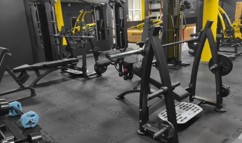 Gymgulv med sorte og gule styrketreningsapparater, flere benkpresser og manualer i treningsstudio, skrå vinkel.