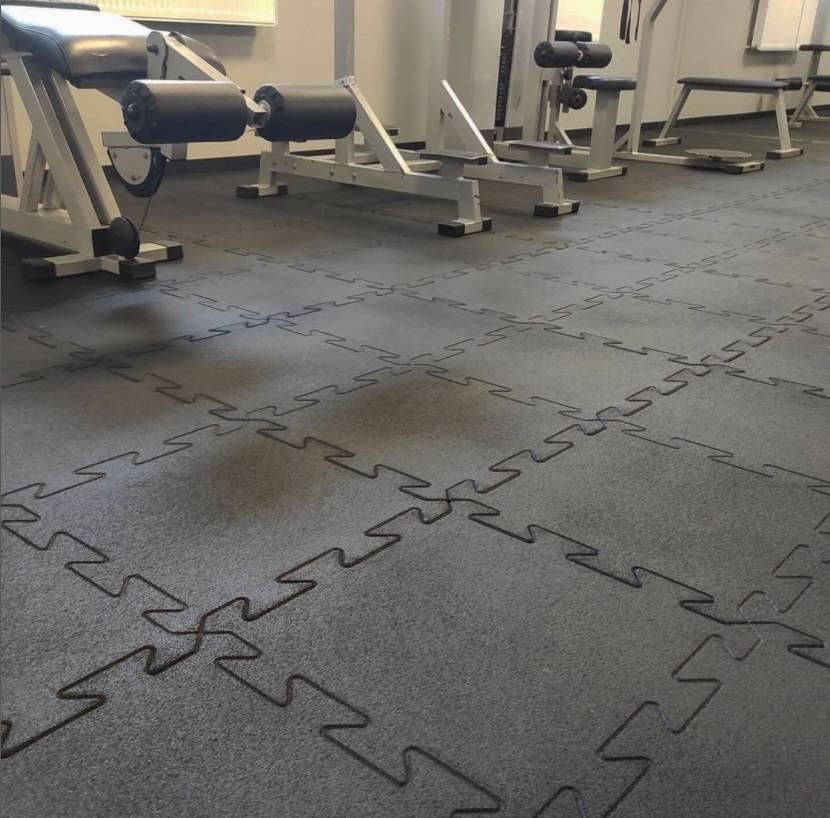 Gymmatte i grått puslespillmønster koblet sammen på gulv i treningsrom, treningsutstyr i bakgrunnen.