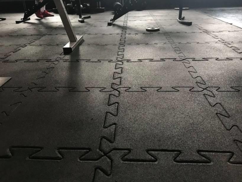 Gummimatten består av sorte puslespill-fliser, lagt ut på gulvet i et hjemmegym med treningsutstyr i bakgrunnen.