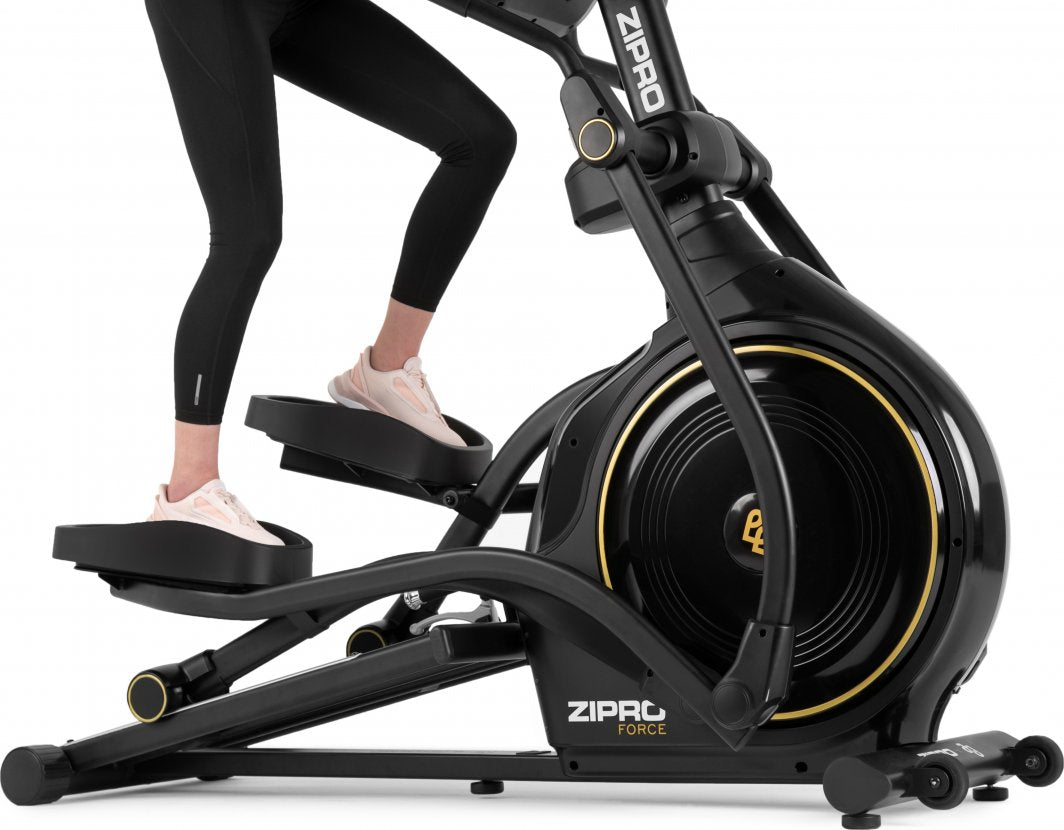 Orbitrek sort ellipsemaskin med gule detaljer, brede fotplater og solid ramme, ben til person i bruk vises.