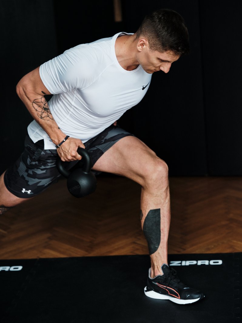 Kettlebells brukes av mann i hvit trøye og svarte shorts som trener roøvelse hjemme med 8 kg vekt.