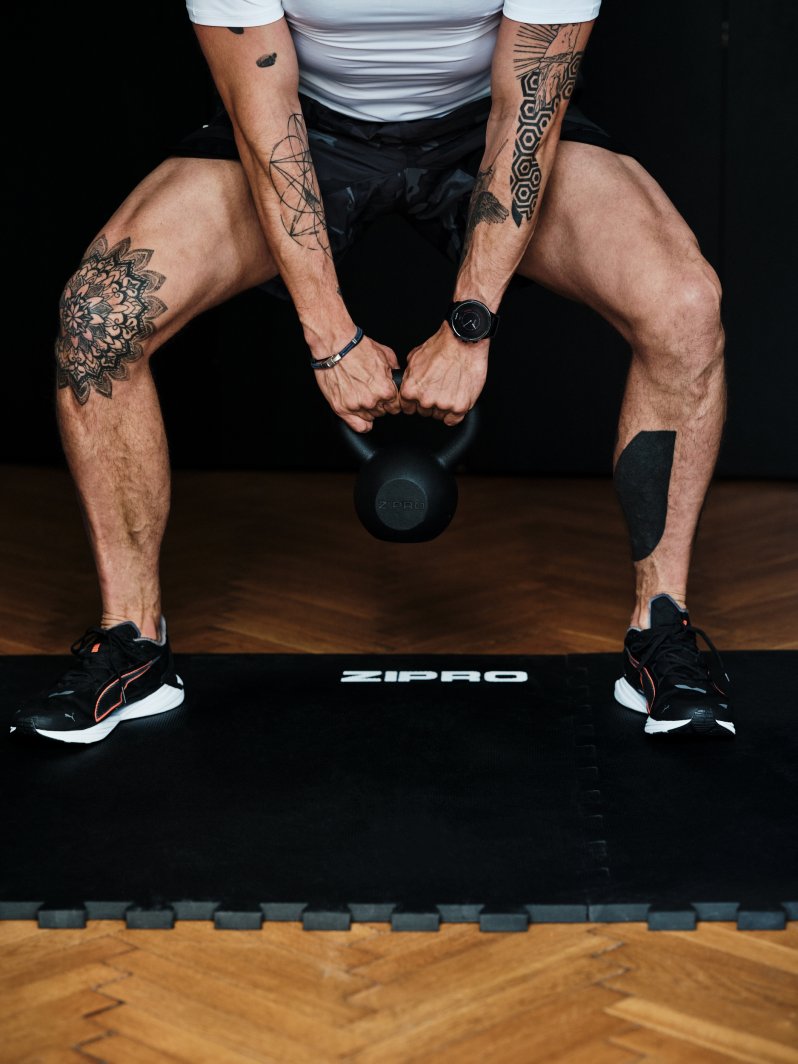 Styrketrening Hjemme med mann som løfter svart kettlebell fra Zipro over svart treningsmatte, synlige tatoveringer.