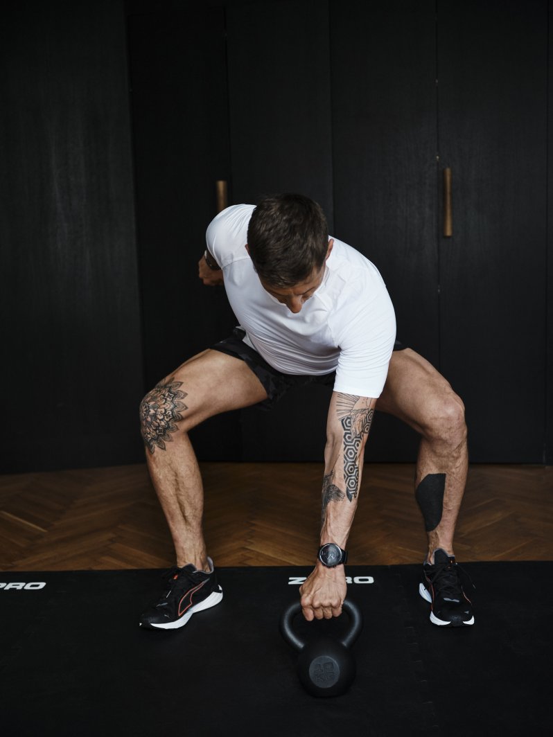 Kettlebell i bruk av mann med hvit t-skjorte og svarte shorts, foran mørke skap i treningsrom.