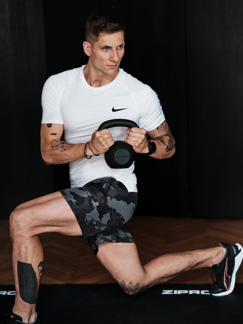 Kettlebells 8kg sort støpejern løftes av mann i hvit T-skjorte og kamuflasjeshorts på treningsmatte.