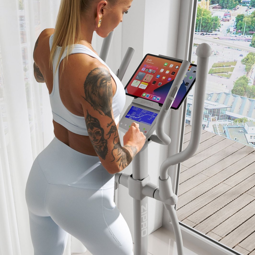 Ellipsemaskin Ned I Vekt lysegrå modell brukt av kvinne i lyst treningstøy, nettbrett og skjerm synlig på konsollen.