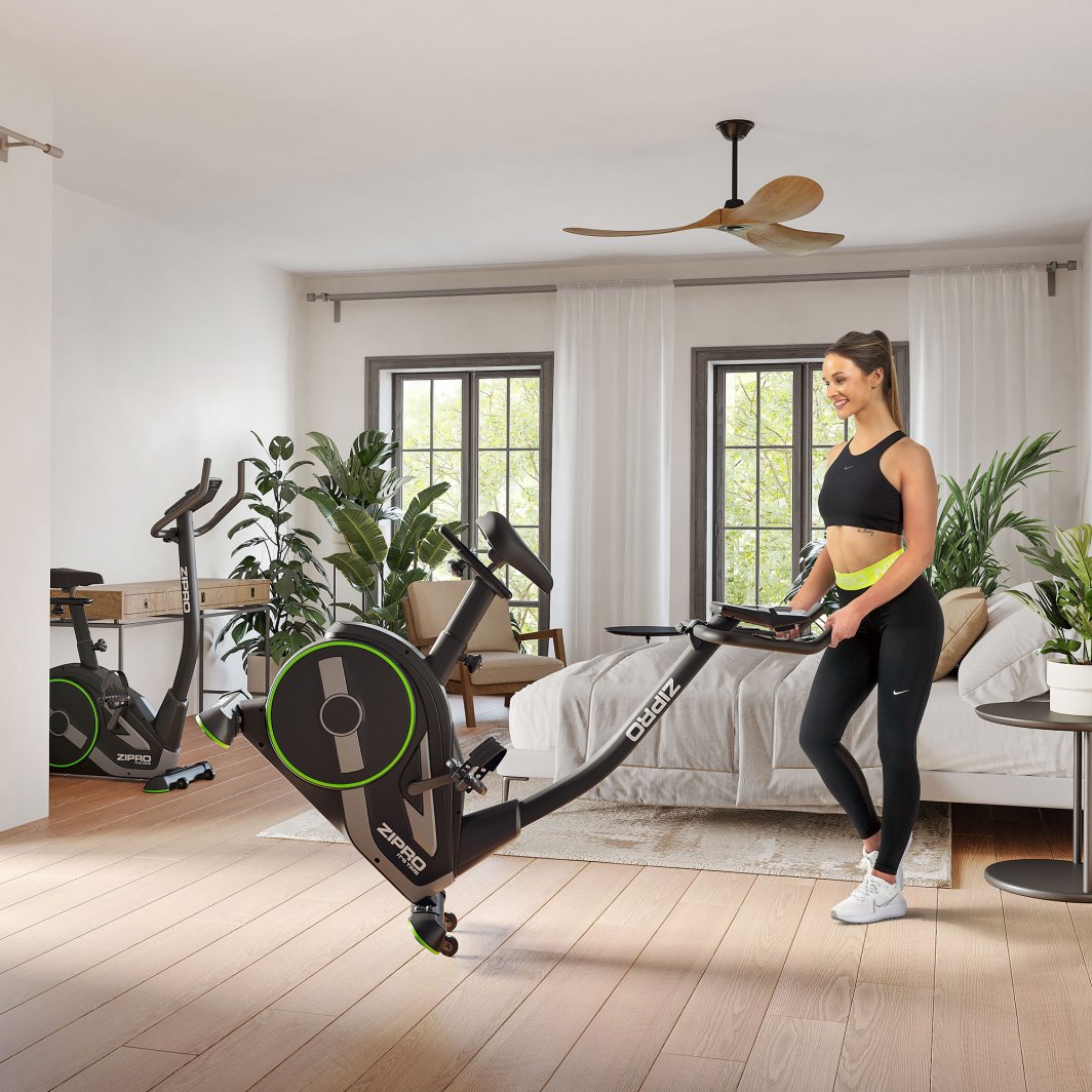 Spinningsykkel i sort med grønne detaljer trilles av kvinne i treningsklær på lyst soverom med planter.