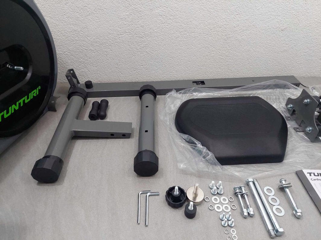 Romaskin Sammenleggbar deler ligger flatt på gulvet, inkludert stålrammer, plastsete, bolter og umbrakonøkler.