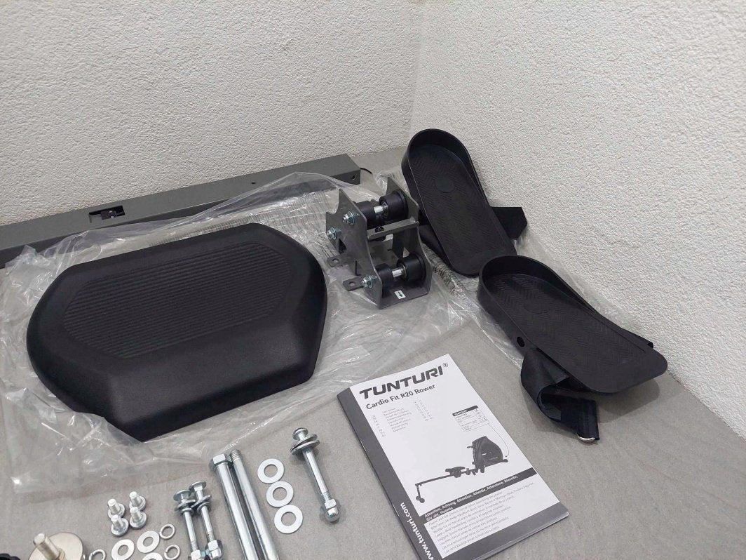 Romaskin med sorte plastdeler, metallbeslag, hjul, pedaler og skruer lagt ut på et bord med bruksanvisning.