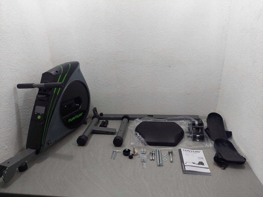 Romaskin Sammenleggbar med svarte og grå deler, instruksjonshefte og monteringsskruer liggende på et bord.