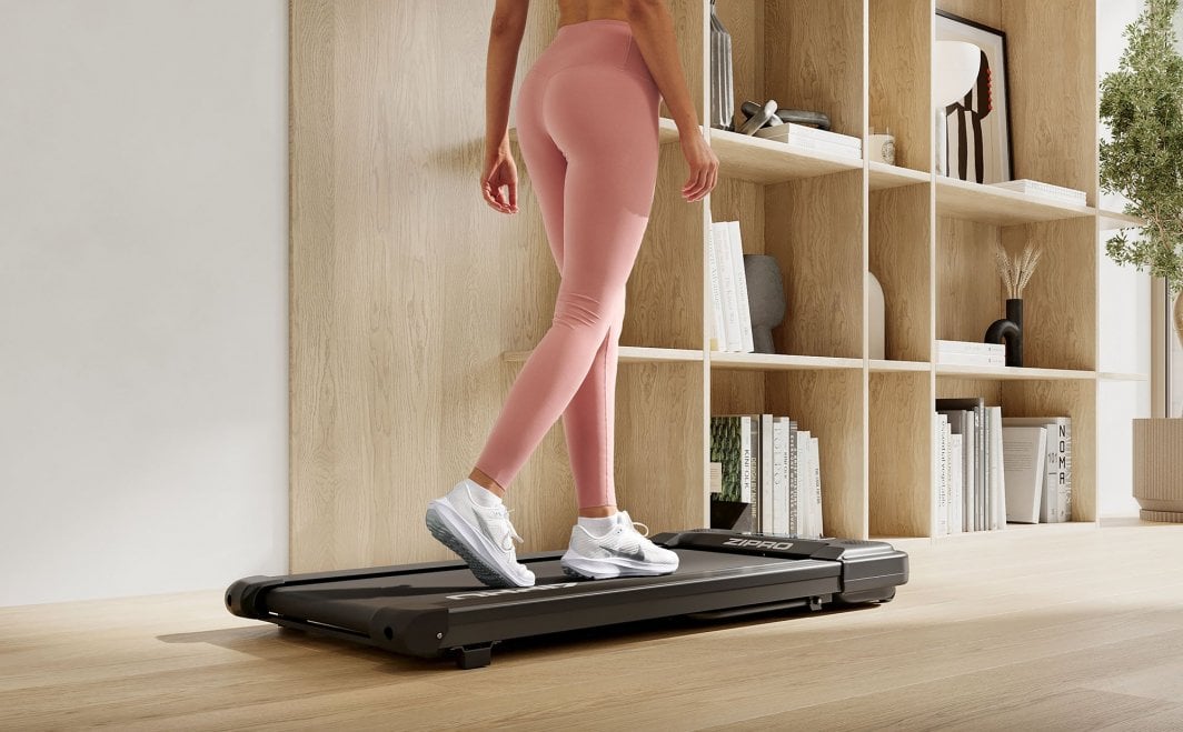 Walking Pad sort tredemølle sett fra siden, en person i rosa treningstights går på den foran en lys bokhylle.