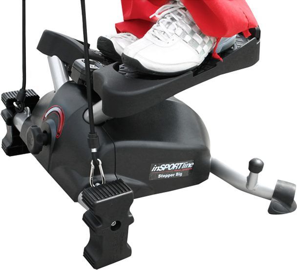Mini Stepper med sorte og grå detaljer, to føtter på pedalene, motstandsbånd foran og ben i røde bukser.
