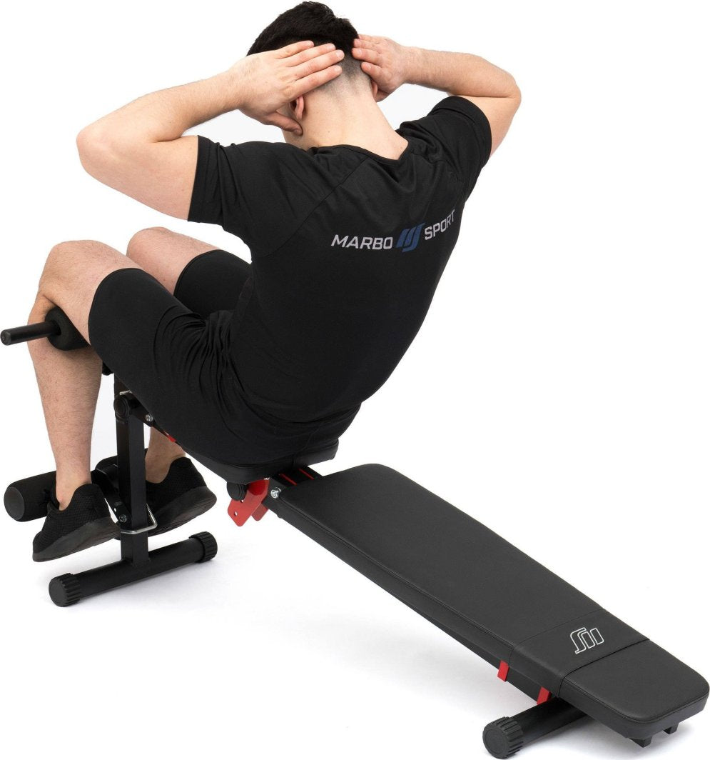 Treningsbenk med svart polstring og metallramme, vist bakfra, person gjør situps med føttene under ruller.