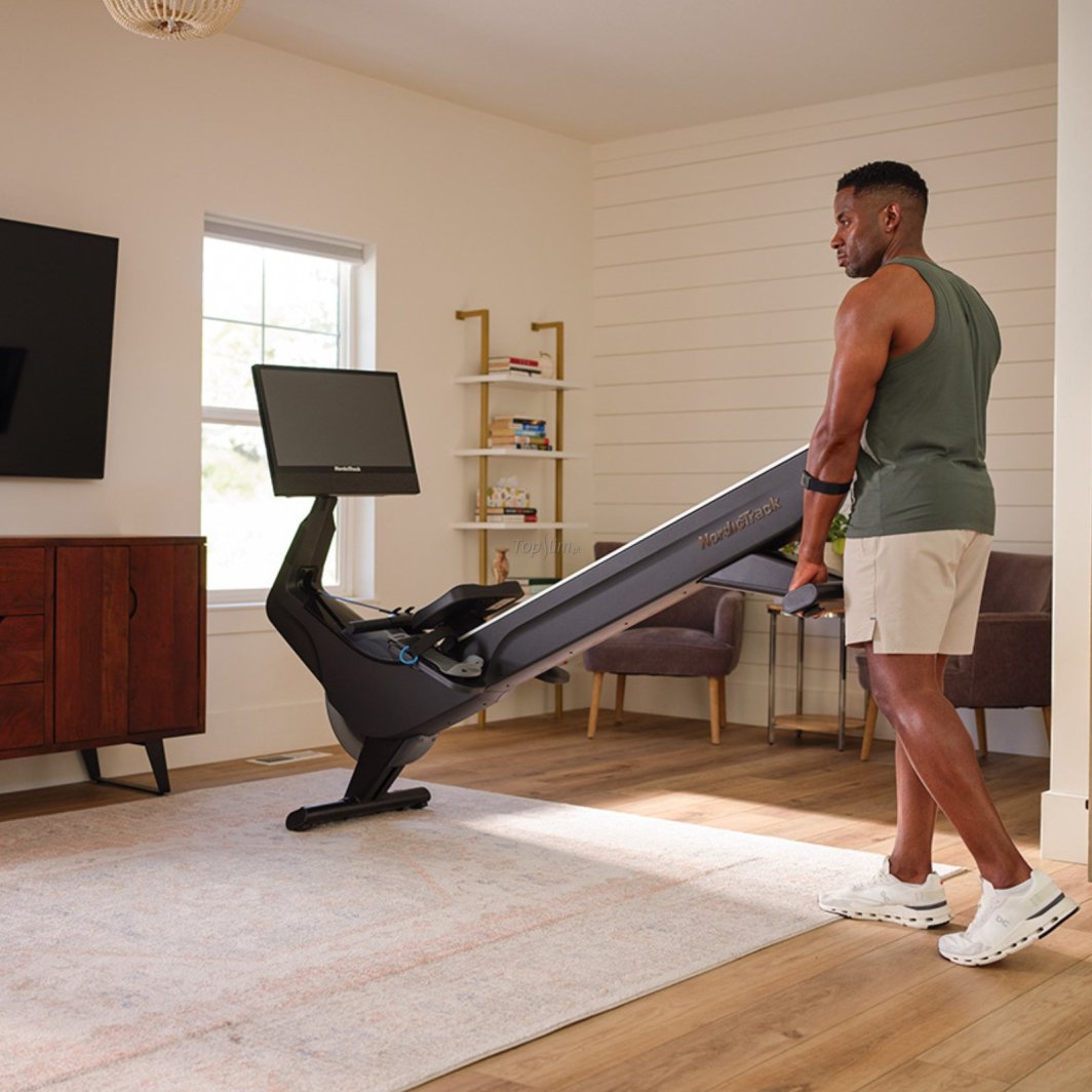 Romaskin med stor skjerm trilles av en mann over lyst teppe i moderne stue, sett fra siden.