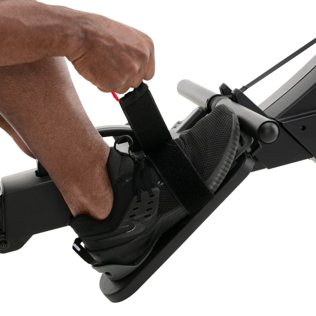 Romaskin Trening med nærbilde av sort sko på fotplate, hånd strammer justerbar reim på treningsmaskinen.