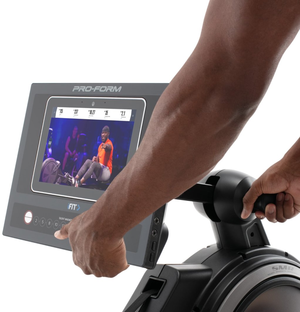 Romaskin Trening med hånd som justerer 10" berøringsskjerm som viser treningsvideo og data på svart, grått apparat.