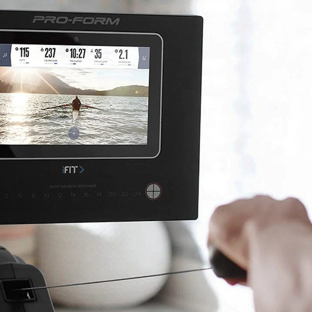 Romaskin med stor digital skjerm som viser treningsprogram, sett fra siden med en hånd på håndtaket.