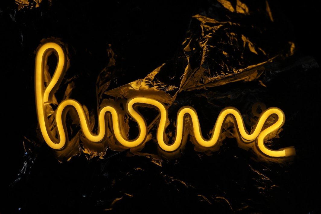 Løpebånd neonskilt HOME i gul utførelse, lyser mot mørk bakgrunn, sett forfra.