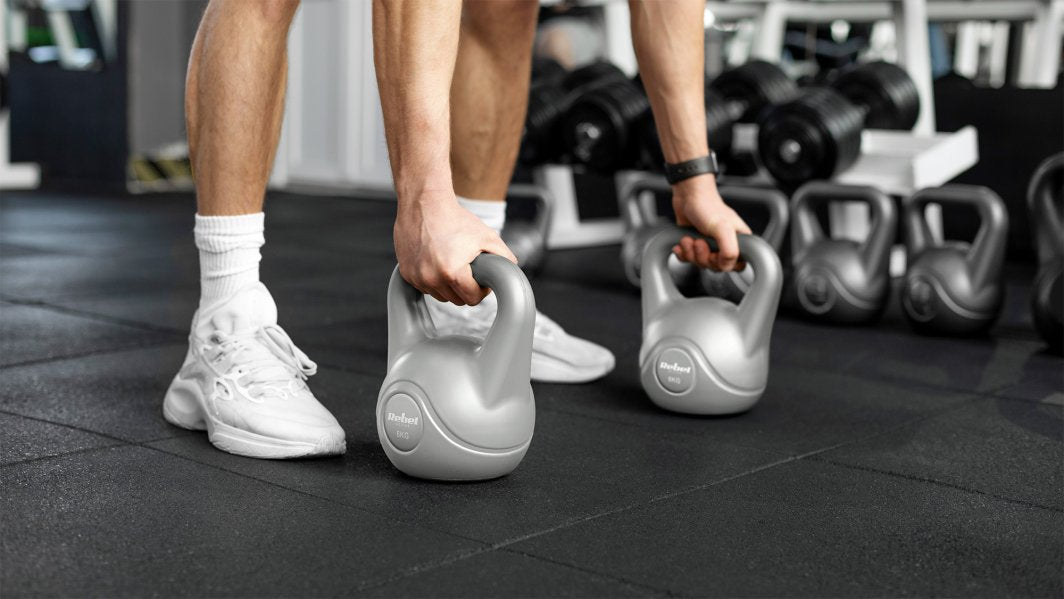 Kettlebell 16 kg løftes av en person med hvite sko i treningsstudio, to grå plastkettlebells med vektmarkering.
