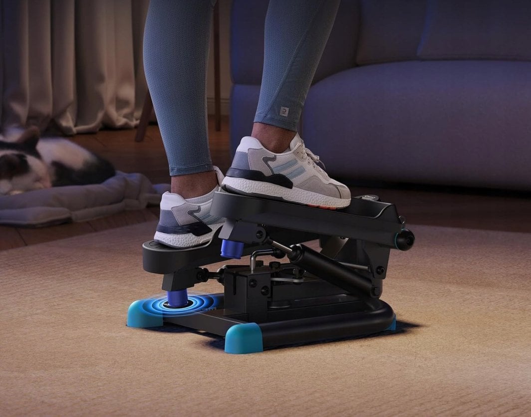 Stepper Trening svart og blå stepmaskin i bruk, to føtter med joggesko, sofa og teppe i bakgrunnen.