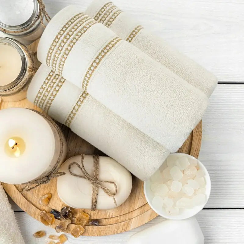 3X Håndkle Amanda, tre kremfargede håndklær med beige border rullet på lyst brett, omgitt av såpe og steiner.