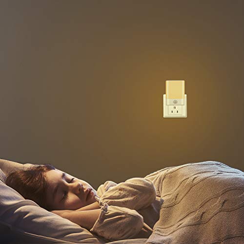 Nattlampe i hvit og gul plast plugget i veggkontakt, lyser varmt over et sovende barn i seng.