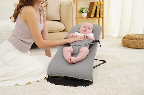 Trekk Til Babybjörn vippestol i grå bomull med baby festet i sele, voksen hånd støtter babyen på magen.