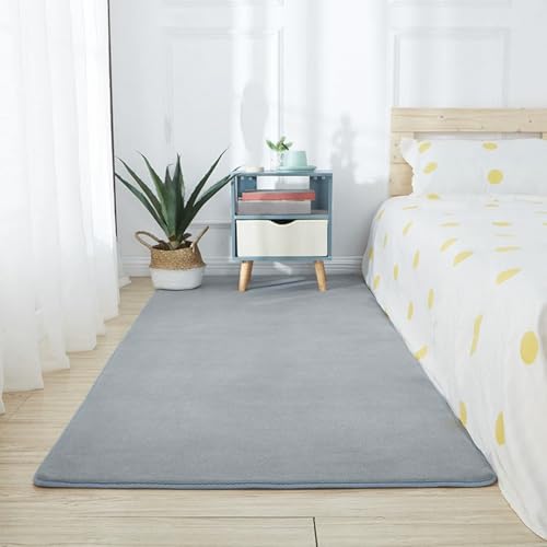 Lekematte Baby i sølvgrått ligger på soveromsgulv ved seng med lyse tekstiler, nattbord, plante og bøker.