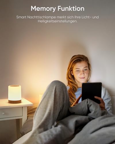 Lampe med hvit skjerm og mørk base lyser på nattbord ved siden av en kvinne som bruker nettbrett i sengen.