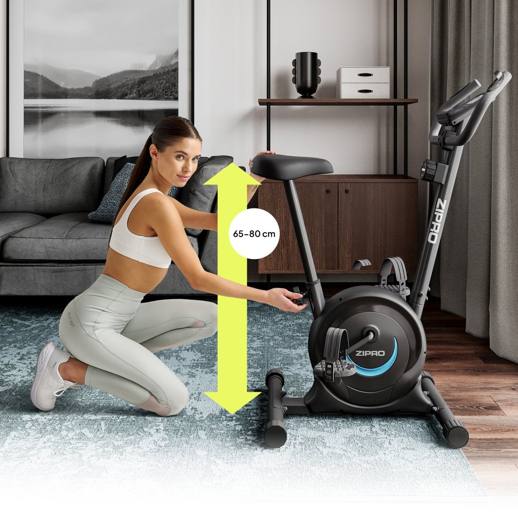 Trimsykkel i sort med blå detaljer, kvinne justerer setet, gul pil viser setehøyde 65–80 cm.