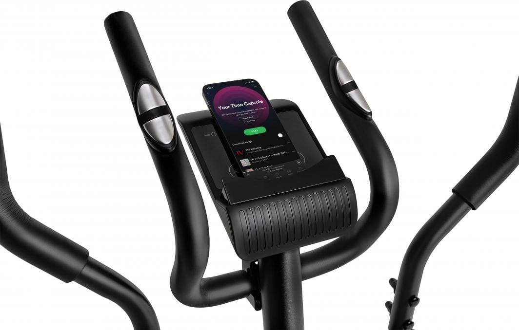 Crosstrainer i sort metall med sølvfargede håndtakssensorer og mobiltelefon i holder foran LCD-displayet.