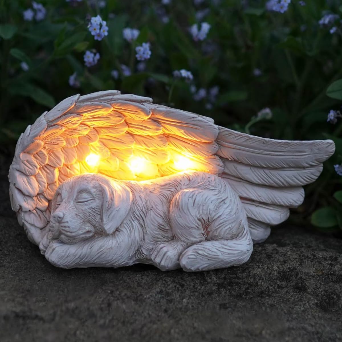 Gravlys formet som sovende beige hund under stor englevinge med varm hvit LED-lys, omgitt av blomster.