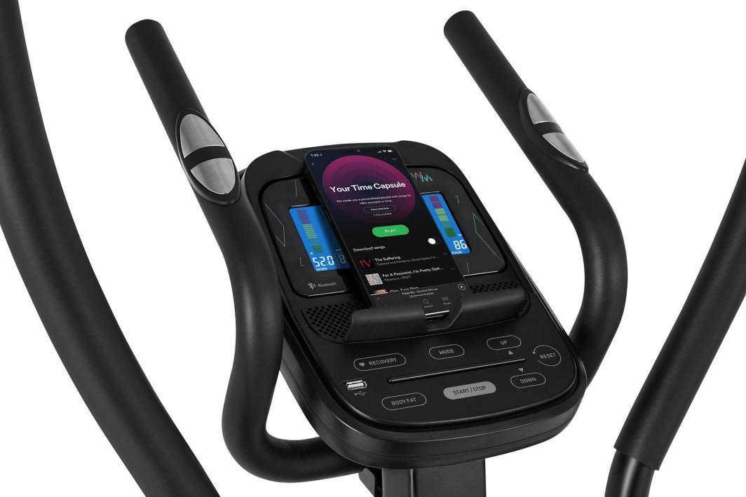 Orbitrek med svarte håndtak, sølvfargede pulssensorer og digitalt display, mobiltelefon i konsollholderen.