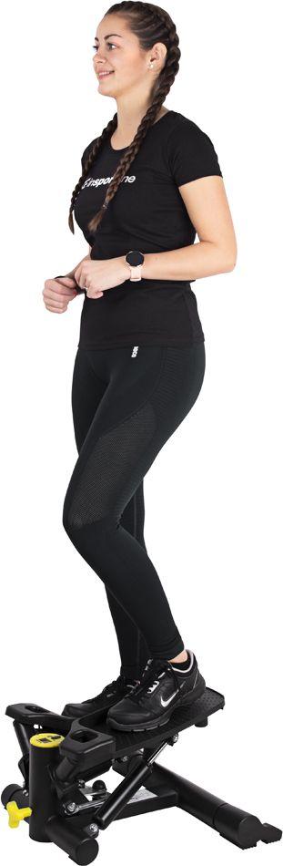 Stepmaskin med vrifunksjon og treningsstrikker brukes av kvinne i treningsklær, gule detaljer vises.