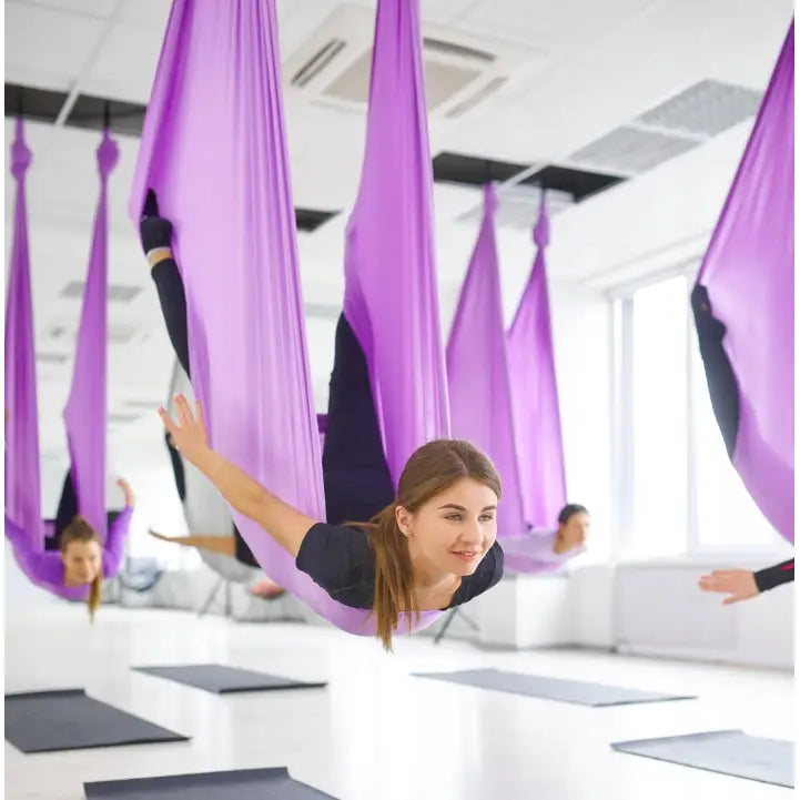 Akrobatisk silkesjerf i lilla, kvinne utfører øvelse i yogaslynge, lyst treningsstudio med store vinduer.