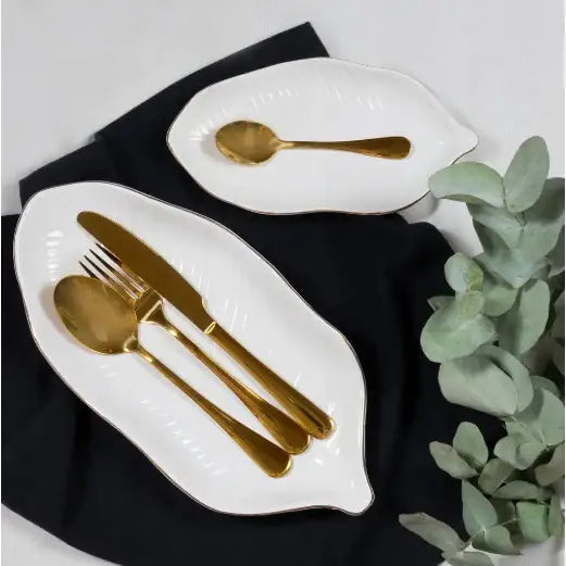 Ambition Enna gullfarget bestikksett, elegant dandert på hvite bladformede tallerkener med grønne blader.