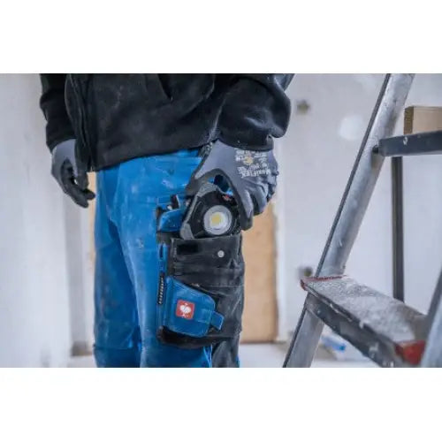 Arbeidslykt LED SH 1000 MA i sort og blå verktøylomme på låret, person ved stige innendørs.