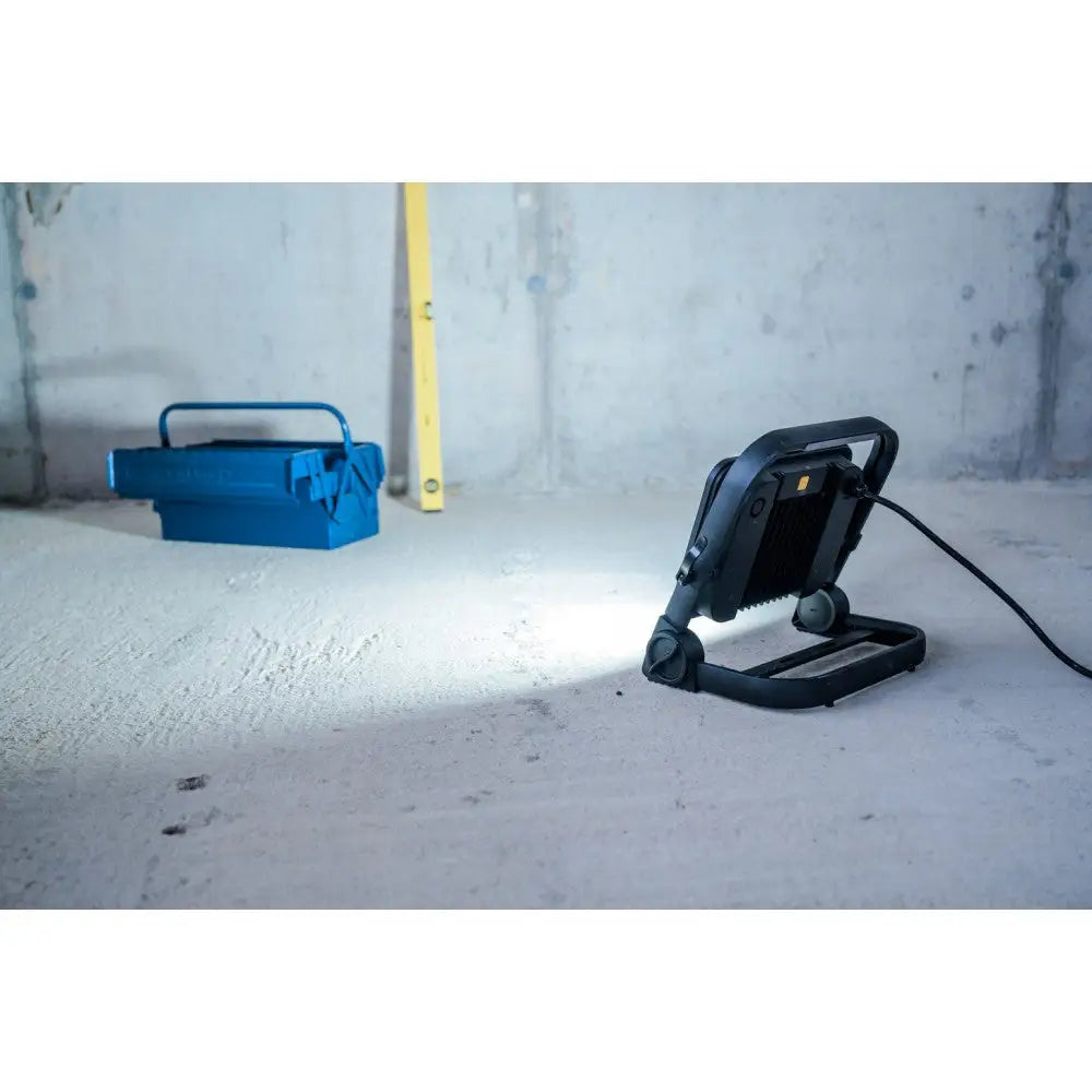 LED arbeidslampe i sort metall, sidevisning på betonggulv med blå verktøykasse og gult vater bak.