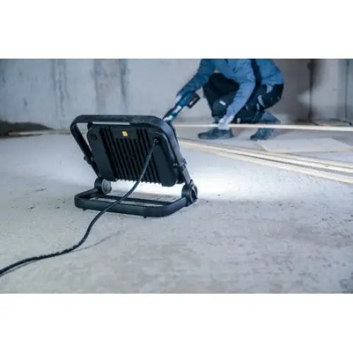 Arbeidslampe LED JARO 14060 M, bakside med tilkoblet ledning, person i verneklær arbeider i bakgrunnen.