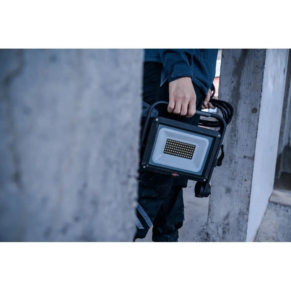 Arbeidslys LED JARO, bæres i hånd med ledningen viklet rundt håndtaket, delvis skjult bak betongsøyle.