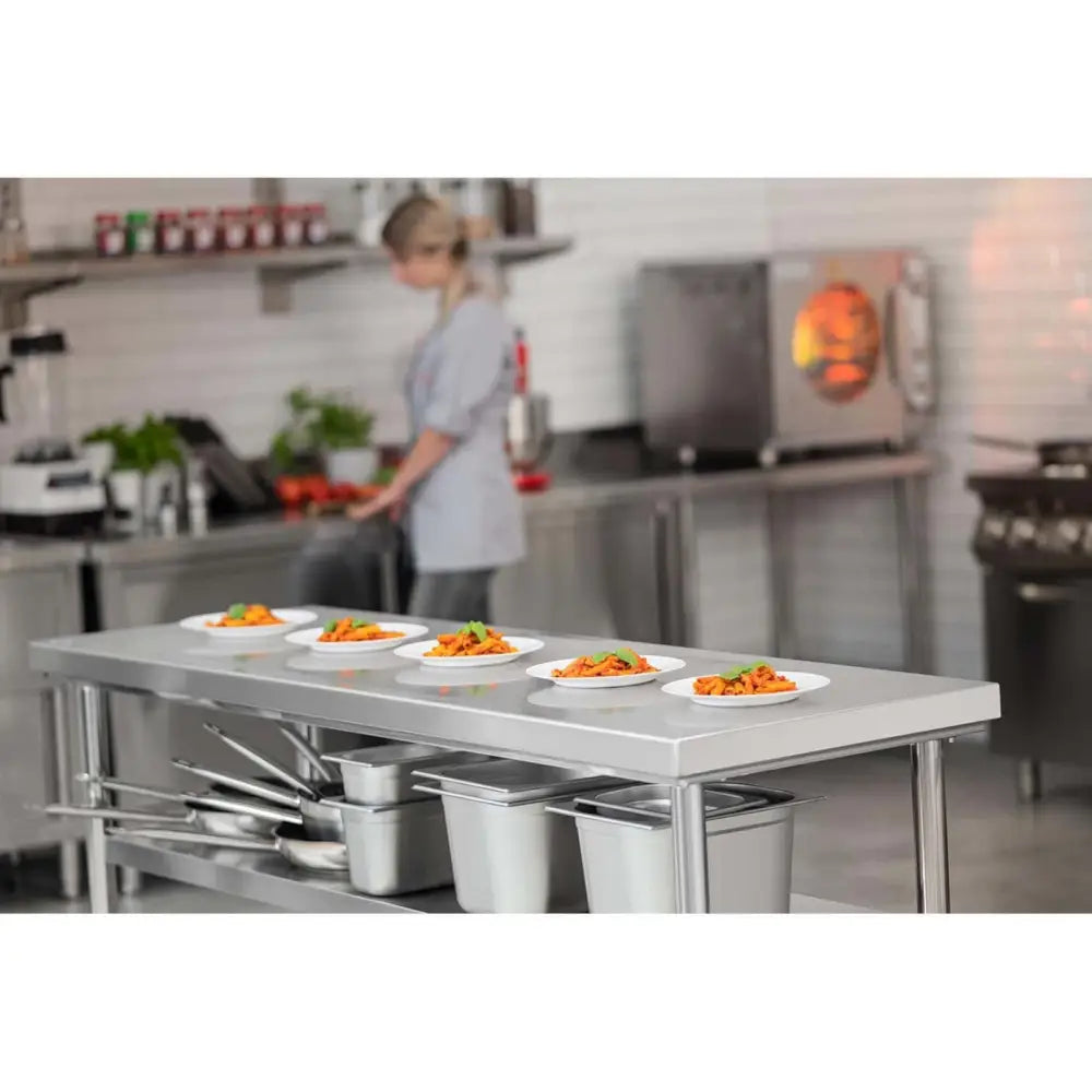 Arbeidsbord sentral med stålplater, sidevisning med fem tallerkener mat og redskaper under i kjøkkenmiljø.