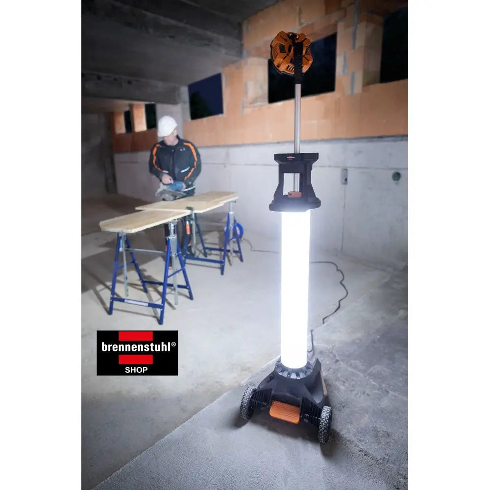 Arbeidslys 360°, stående på hjul med sort og oransje base, lyser opp byggeplass med arbeider i bakgrunnen.