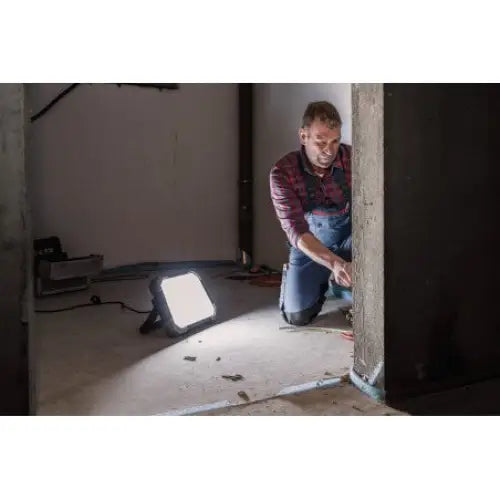 LED arbeidslys DINORA, plassert på gulv og lyser opp et uferdig rom mens en mann arbeider på gulvet.