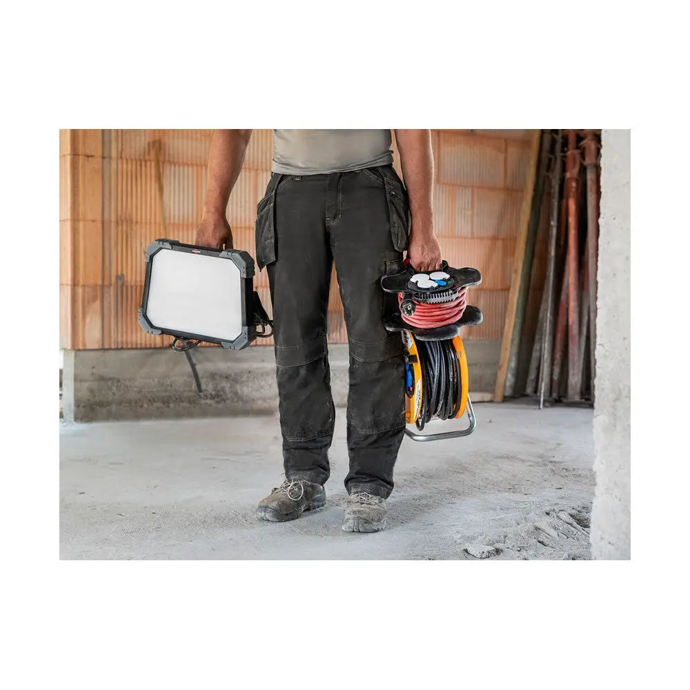 Arbeidslys LED DINORA, bæres av person med arbeidsklær og støvete sko, sammen med kabeltrommel innendørs.