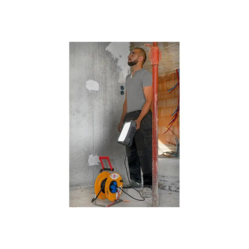 Arbeidslys LED DINORA, holdt av person foran betongvegg, gul kabeltrommel og byggeplass i bakgrunnen.