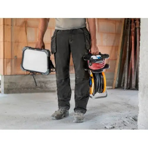 LED arbeidslys DINORA, holdes av person i arbeidsklær sammen med kabeltrommel på byggeplass med rå vegger.