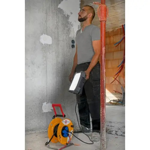 Arbeidslys LED DINORA holdes av håndverker på byggeplass, gul kabeltrommel synlig foran på betonggulv.